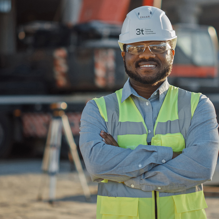 A smiling construction worker, donning a hard hat and safety vest, stands confidently with arms crossed at a construction site. Surrounded by equipment, he embodies the impact of rigorous training in ensuring a safe and efficient work environment.