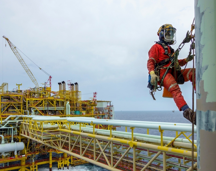 Un trabajador con equipo de seguridad, demostrando una formación experta, está suspendido de cuerdas inspeccionando una estructura vertical en una plataforma petrolífera en alta mar, con el océano y la maquinaria de la plataforma visibles en el fondo.