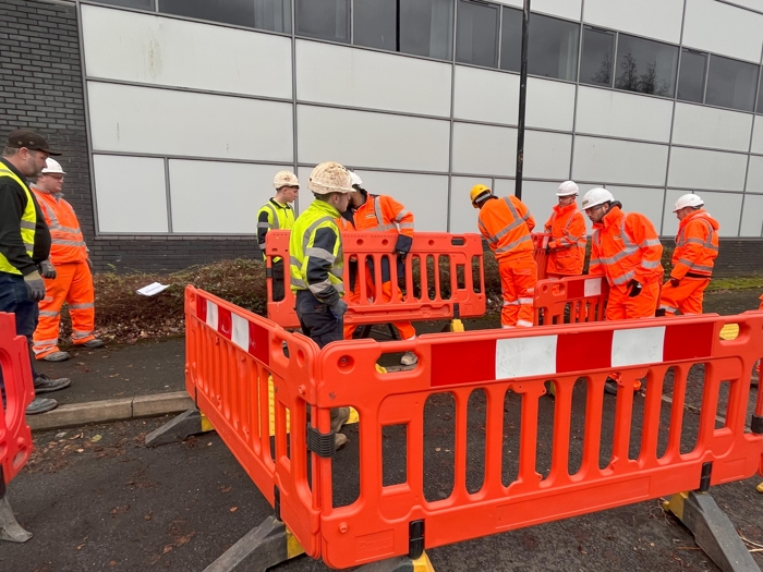 Construction workers in safety gear, undergoing training, stand near orange barriers on a road beside a building.