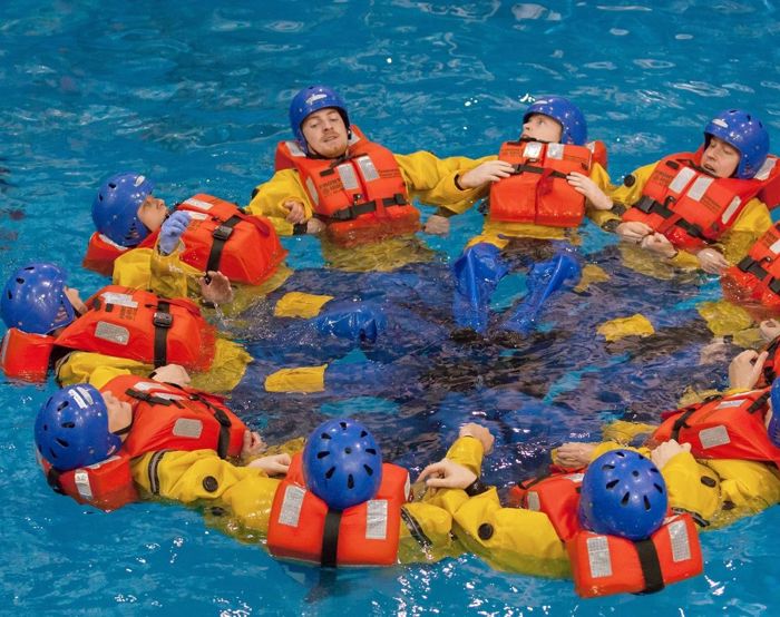 Un grupo de personas con chalecos salvavidas y cascos naranjas forman un círculo mientras flotan en una piscina, participando en un ejercicio de entrenamiento.