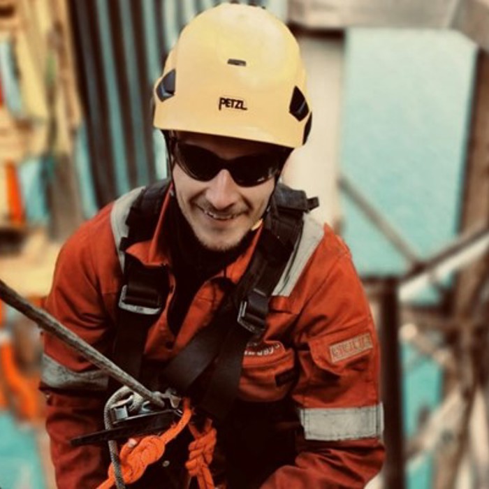 Un trabajador con casco amarillo y equipo de seguridad está escalando una estructura con cuerdas, con una gran masa de agua visible al fondo.