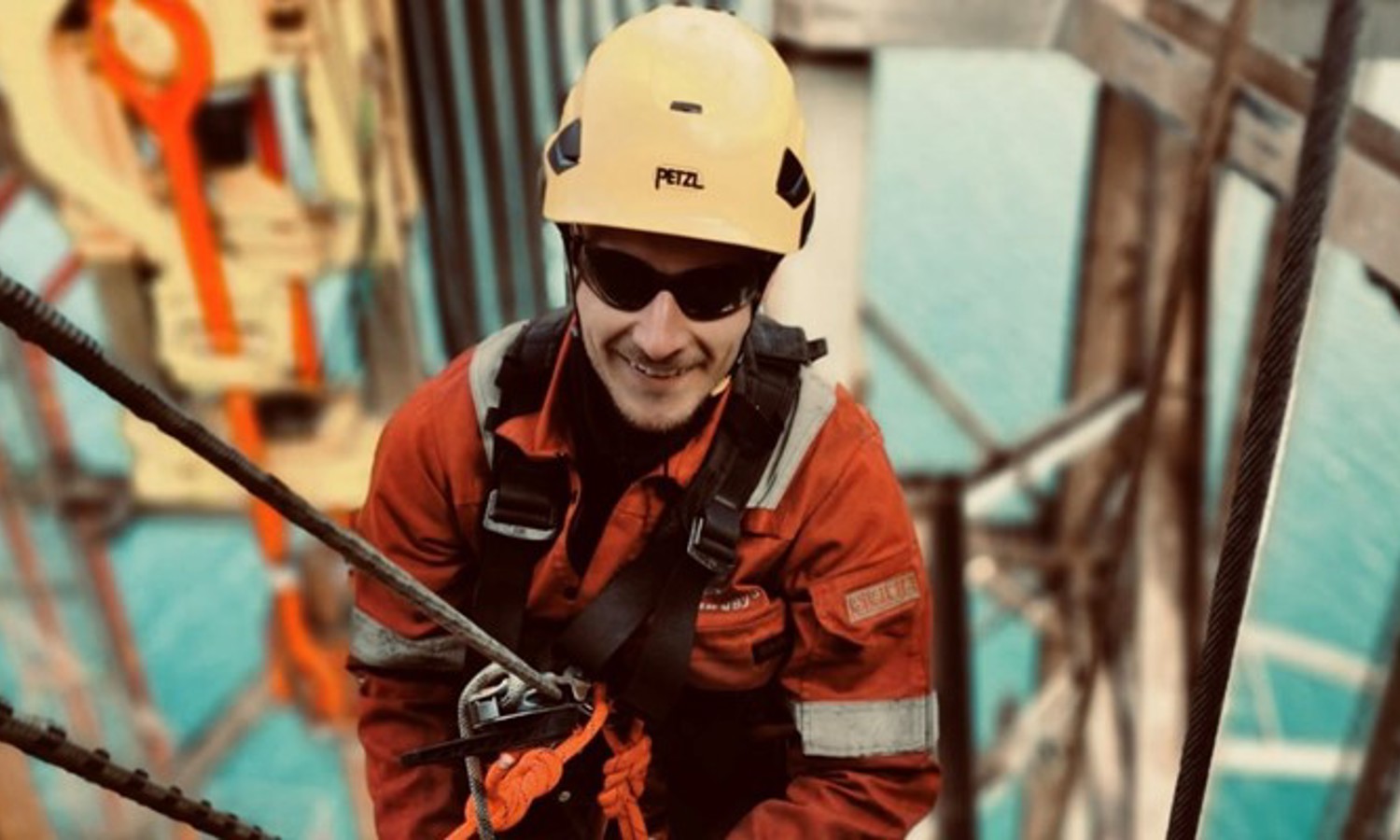 Un trabajador con casco amarillo y equipo de seguridad está escalando una estructura con cuerdas, con una gran masa de agua visible al fondo.