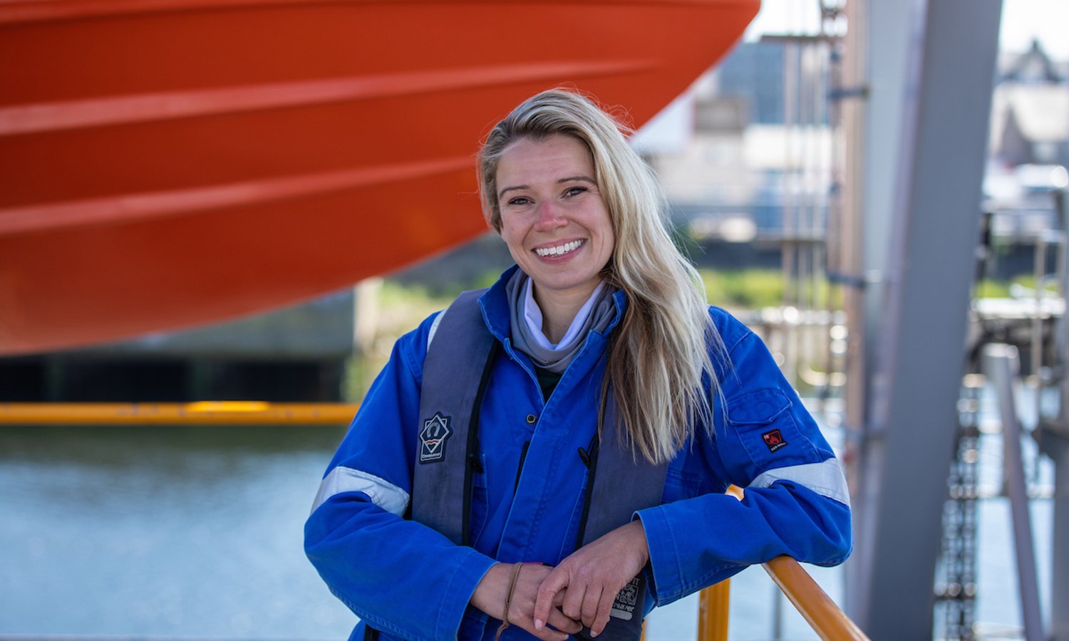 A woman in a blue work uniform and life vest stands smiling outdoors, showcasing her training near a large orange boat.