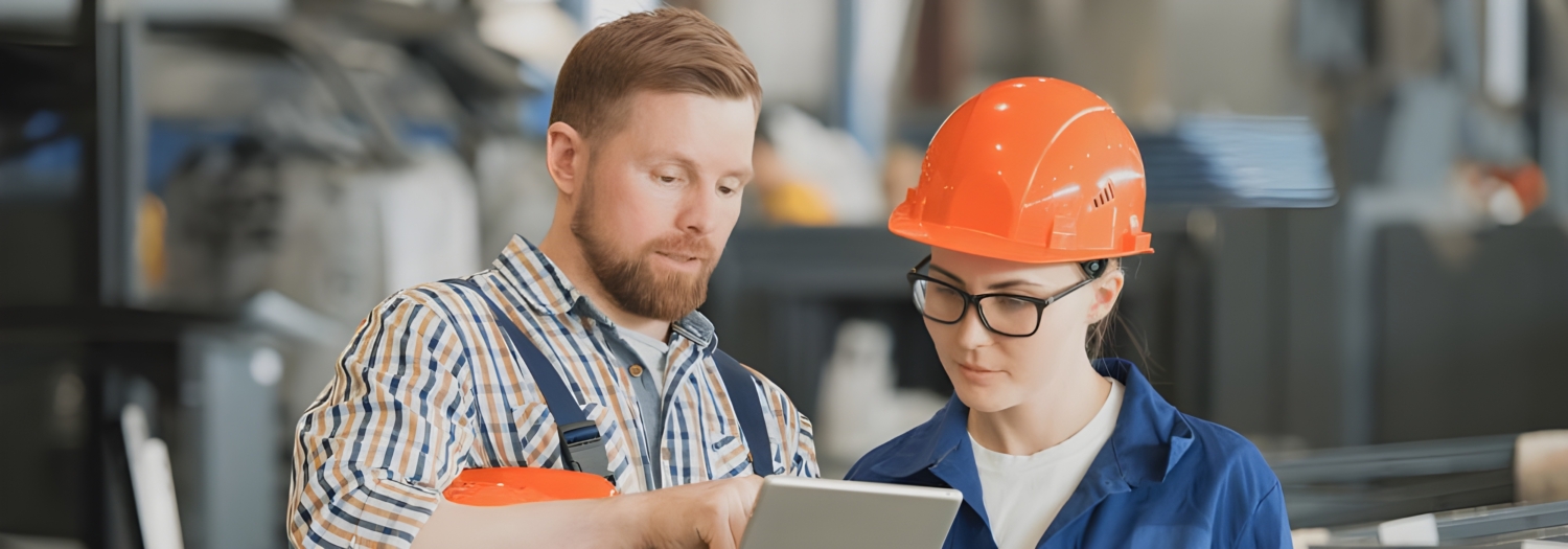 Two workers in a warehouse, one wearing a plaid shirt and the other in a blue uniform with an orange hard hat, are reviewing training information on a tablet.