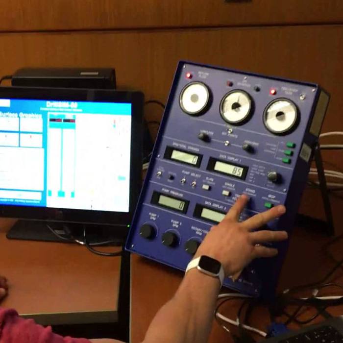 A person operates complex electronic equipment with dials and digital displays at a work station, featuring multiple screens and control panels.
