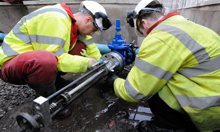 Two workers in high-visibility jackets and hard hats are meticulously inspecting and fixing a blue pipe joint in a construction area, demonstrating their training expertise.