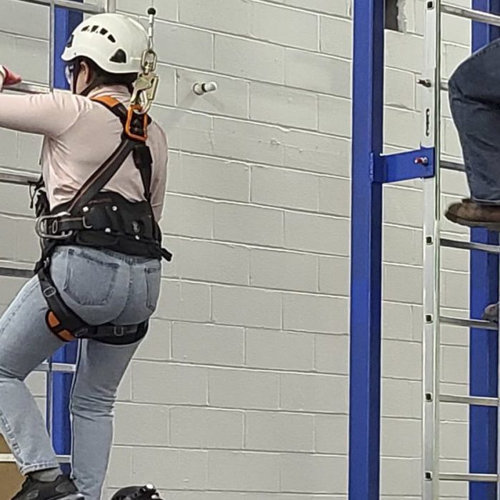 Two individuals in safety gear and helmets are climbing vertical ladders inside an industrial training facility.