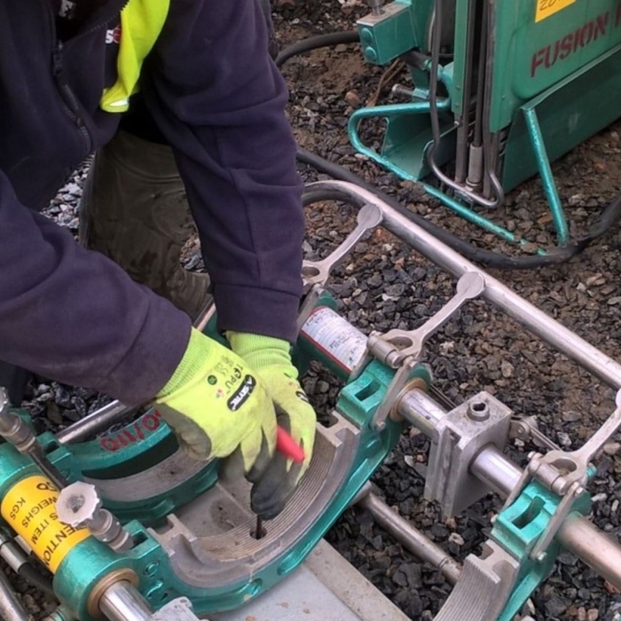 A person wearing gloves and a safety vest, part of a GNO apprenticeship program, uses a tool to adjust a green fusion machine on a construction site with gravel on the ground.