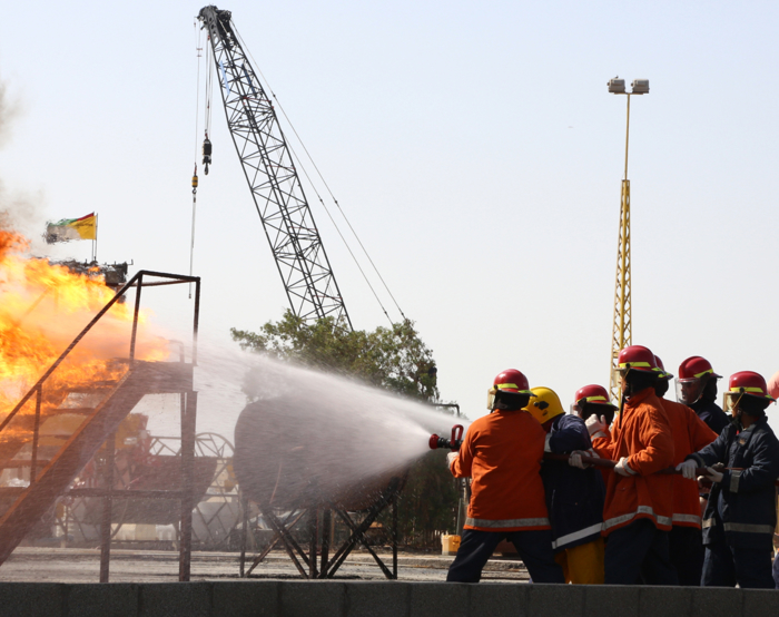 Bomberos con uniformes naranjas y cascos rojos utilizan una manguera para rociar agua sobre un gran incendio industrial con humo espeso. Al fondo se ve una grúa y una bandera.
