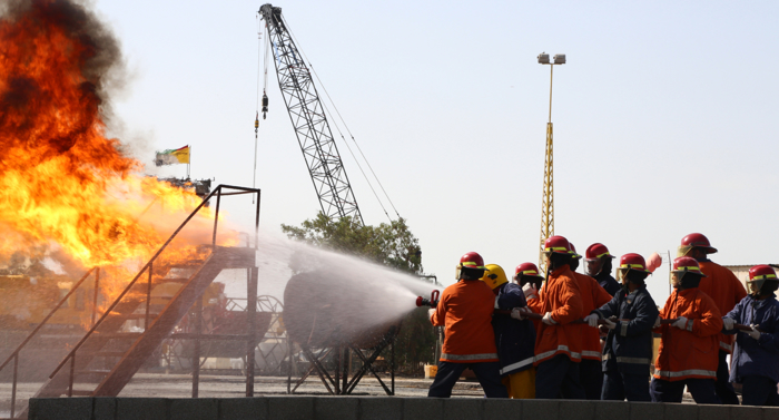 Bomberos con uniformes naranjas y cascos rojos utilizan una manguera para rociar agua sobre un gran incendio industrial con humo espeso. Al fondo se ve una grúa y una bandera.