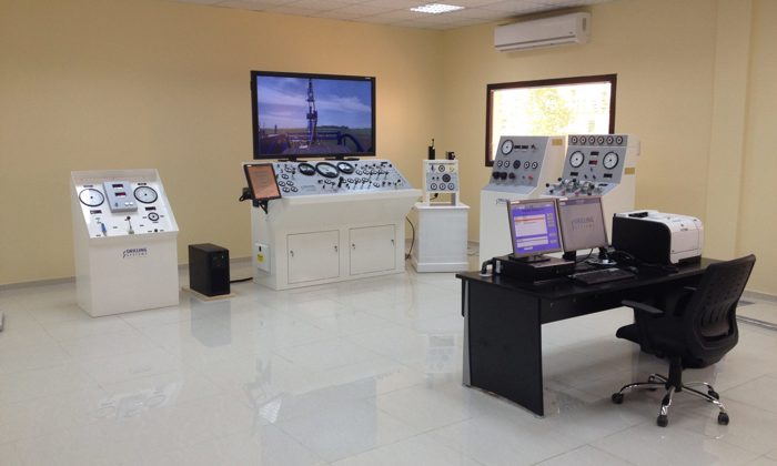 A control room with multiple control panels, a desk with computer monitors, various gauges, and a large screen on the wall displaying an image of an industrial setup.