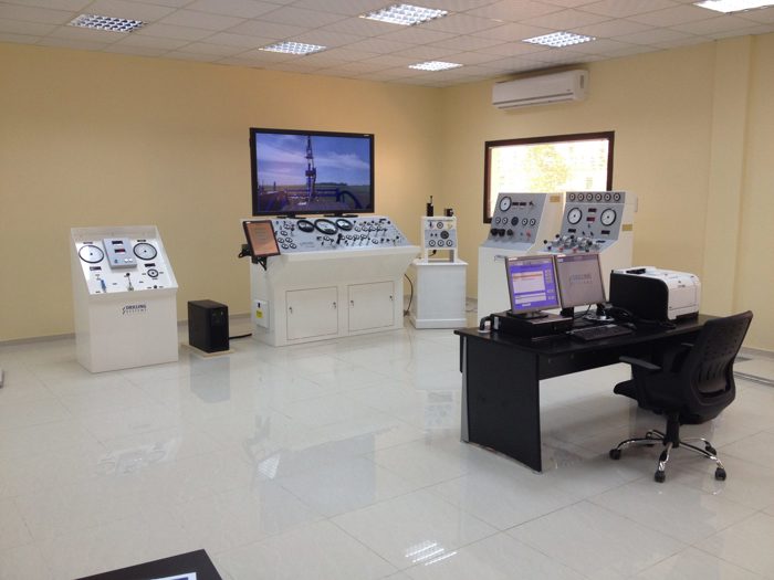 Una sala de control con múltiples paneles de control, un escritorio con monitores de ordenador, varios medidores y una gran pantalla en la pared que muestra la imagen de una instalación industrial.