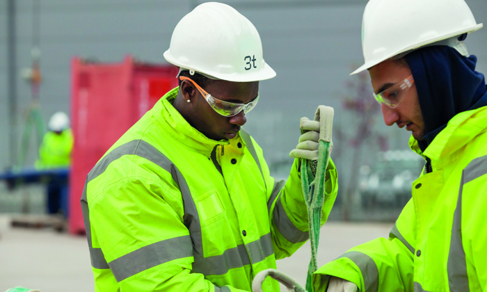 Two workers wearing high-visibility jackets and safety helmets are undergoing training as they skillfully handle a rope or strap outdoors.