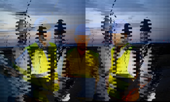 Tres trabajadores con equipo de seguridad y casco reciben formación mientras examinan un ordenador portátil cerca de unos aerogeneradores al anochecer.